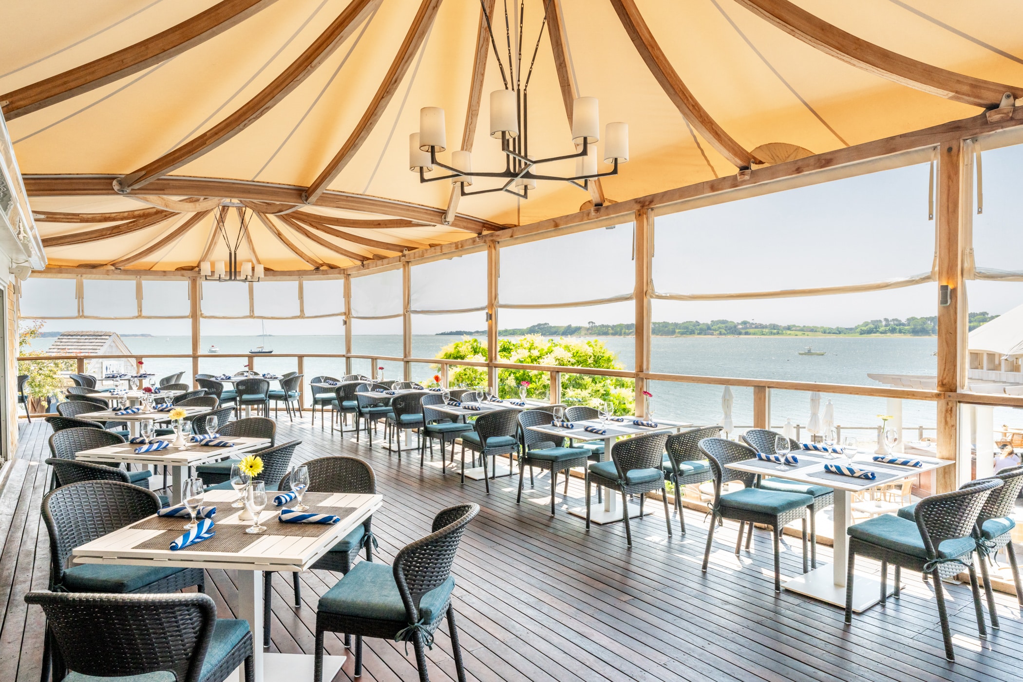 Outdoor restaurant patio with neatly set tables and chairs under a canopy, overlooking a calm body of water on a clear day at the inviting Outer Bar & Grille.