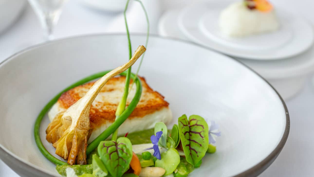 A plated gourmet dish featuring a seared white fish fillet, green vegetables, edible flowers, and a delicate garnish, served in a white bowl on a white tablecloth—perfect for elegant dining accommodations.