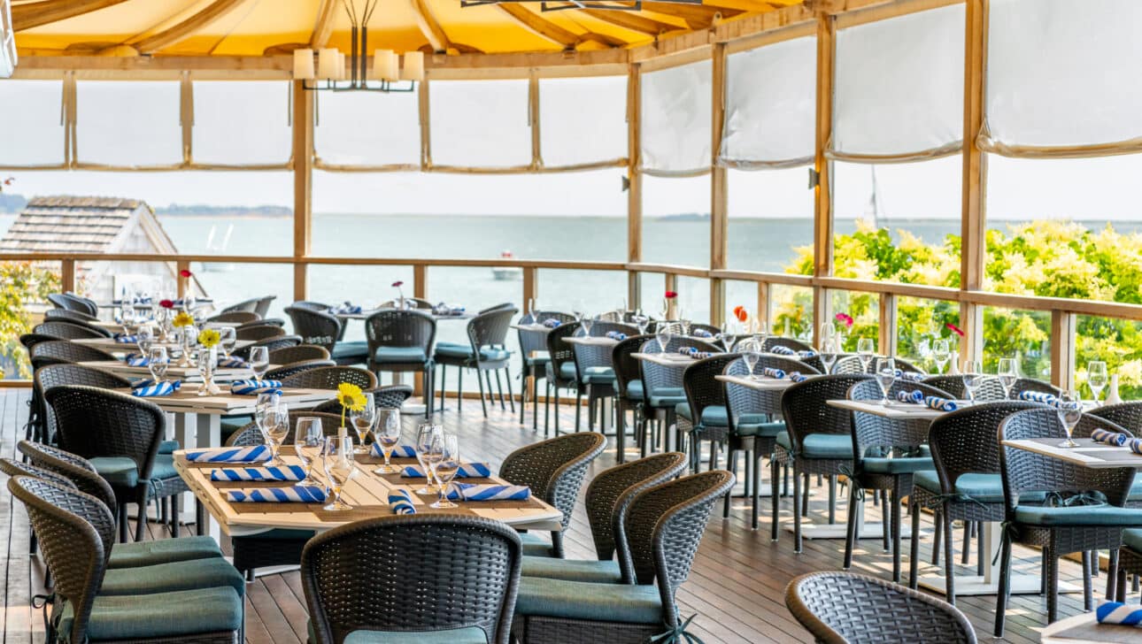 Outdoor restaurant patio with neatly arranged tables and chairs under a canopy, set with glassware and napkins, overlooking a body of water.