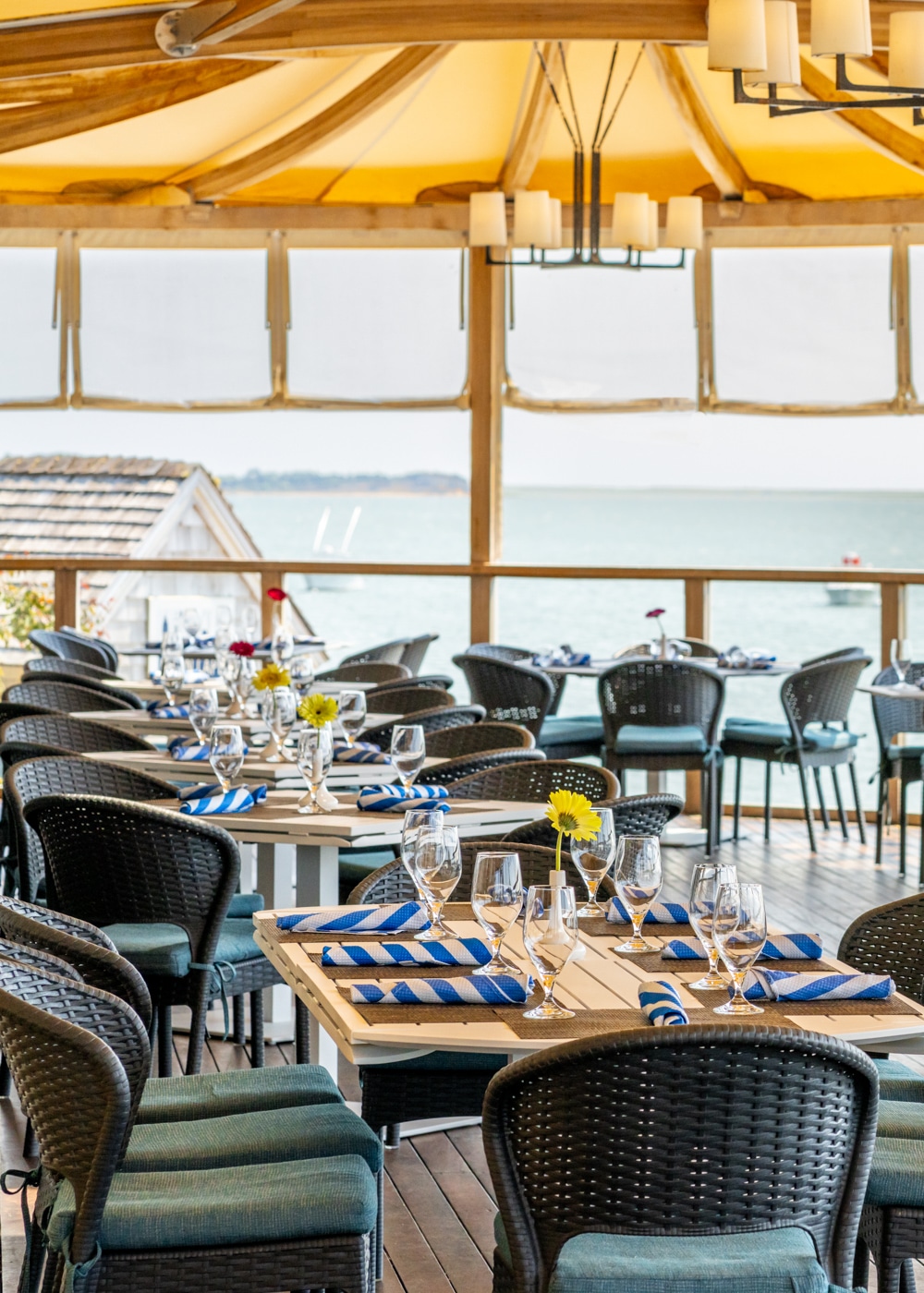 Outdoor restaurant patio at Wequassett Resort with neatly set tables, black wicker chairs, blue-striped napkins, glasses, and yellow flower centerpieces overlooking the waterfront—perfect for relaxing after a round at the golf club.