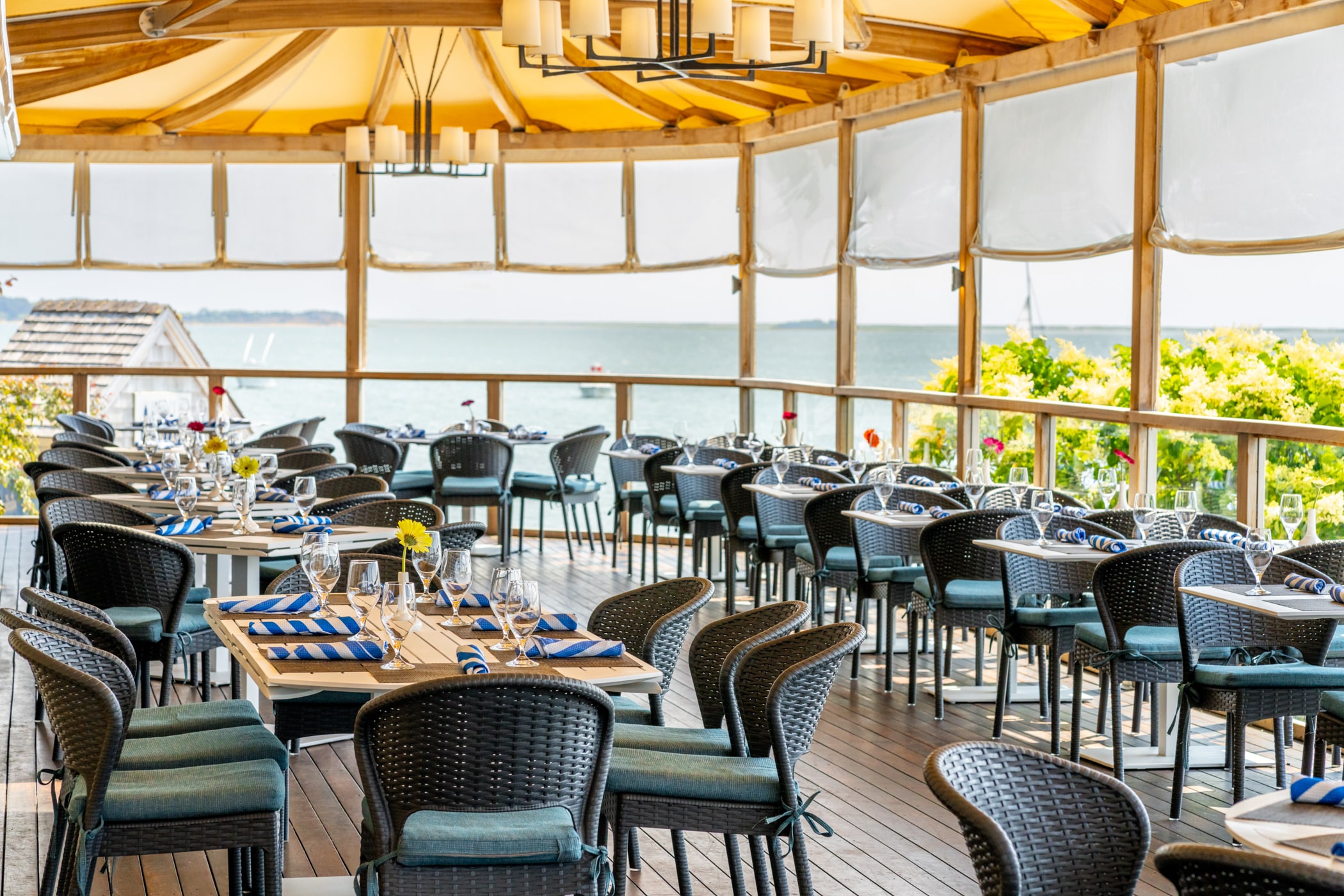 Outdoor restaurant dining area with wicker chairs, set tables, and a yellow canopy at Wequassett Resort, overlooking water and lush greenery in the background.