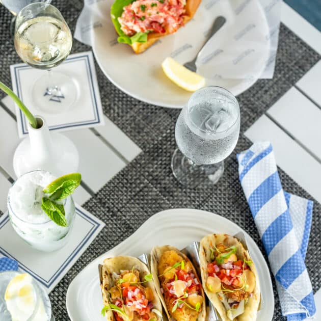 Three soft tacos with toppings on a white plate, a lobster roll on a separate plate, drinks with lemon and mint, and a striped napkin on a table.