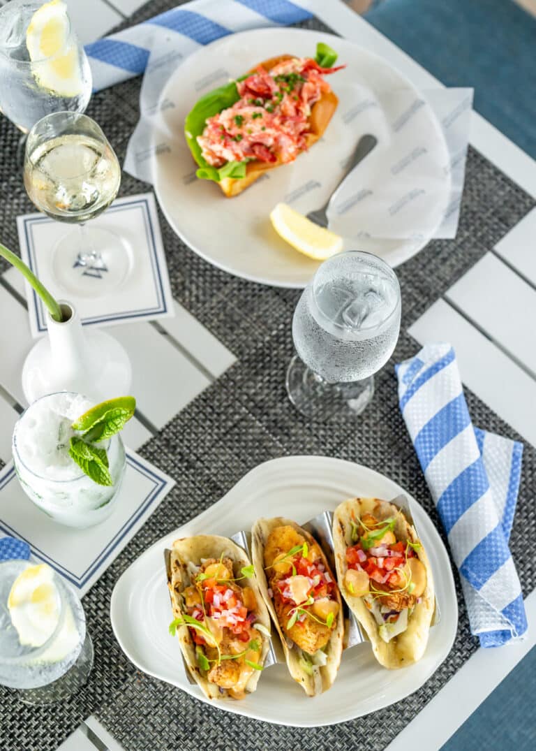 Three soft tacos with toppings on a white plate, a lobster roll on a separate plate, drinks with lemon and mint, and a striped napkin on a table.