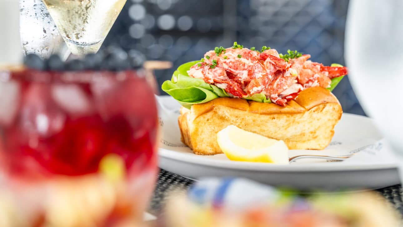 A lobster roll on a plate with lettuce and a lemon wedge, with a glass of white wine and a red beverage in the foreground.