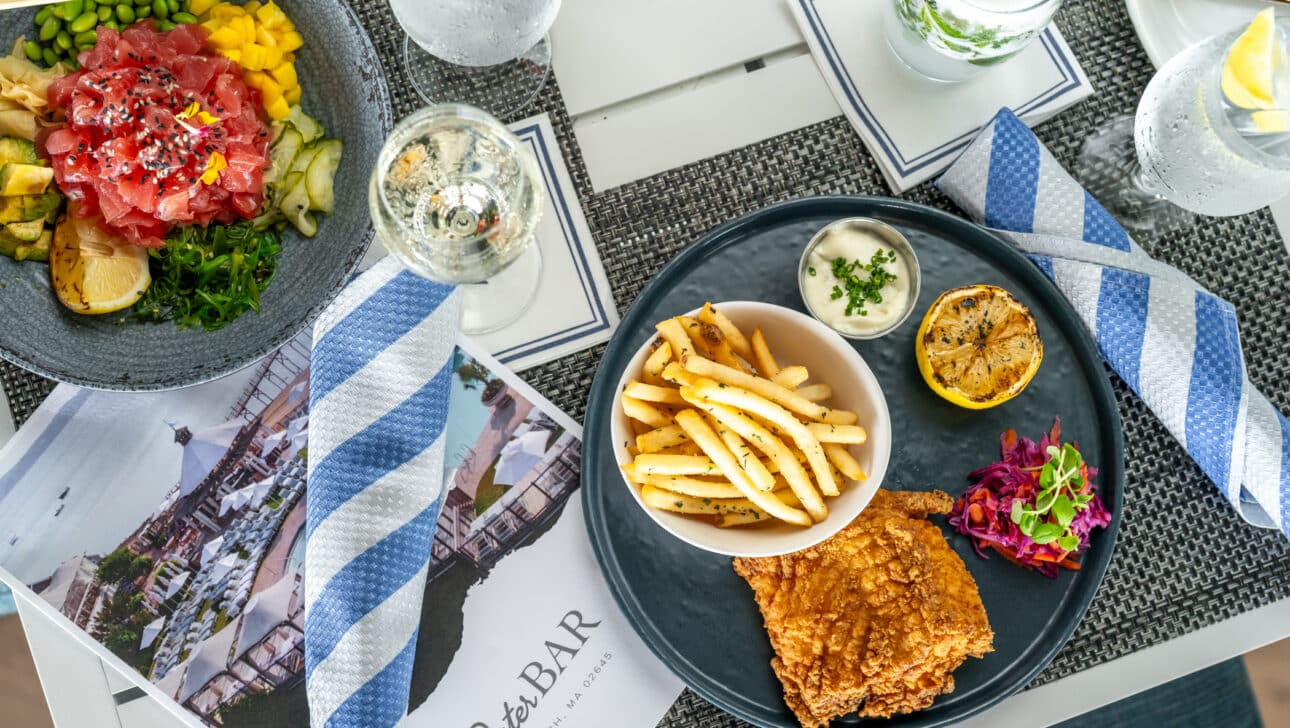 Overhead view of a meal at Wequassett Resort with fried fish, French fries, coleslaw, and a grilled lemon, next to a plate of poke salad and two glasses of water on the table.