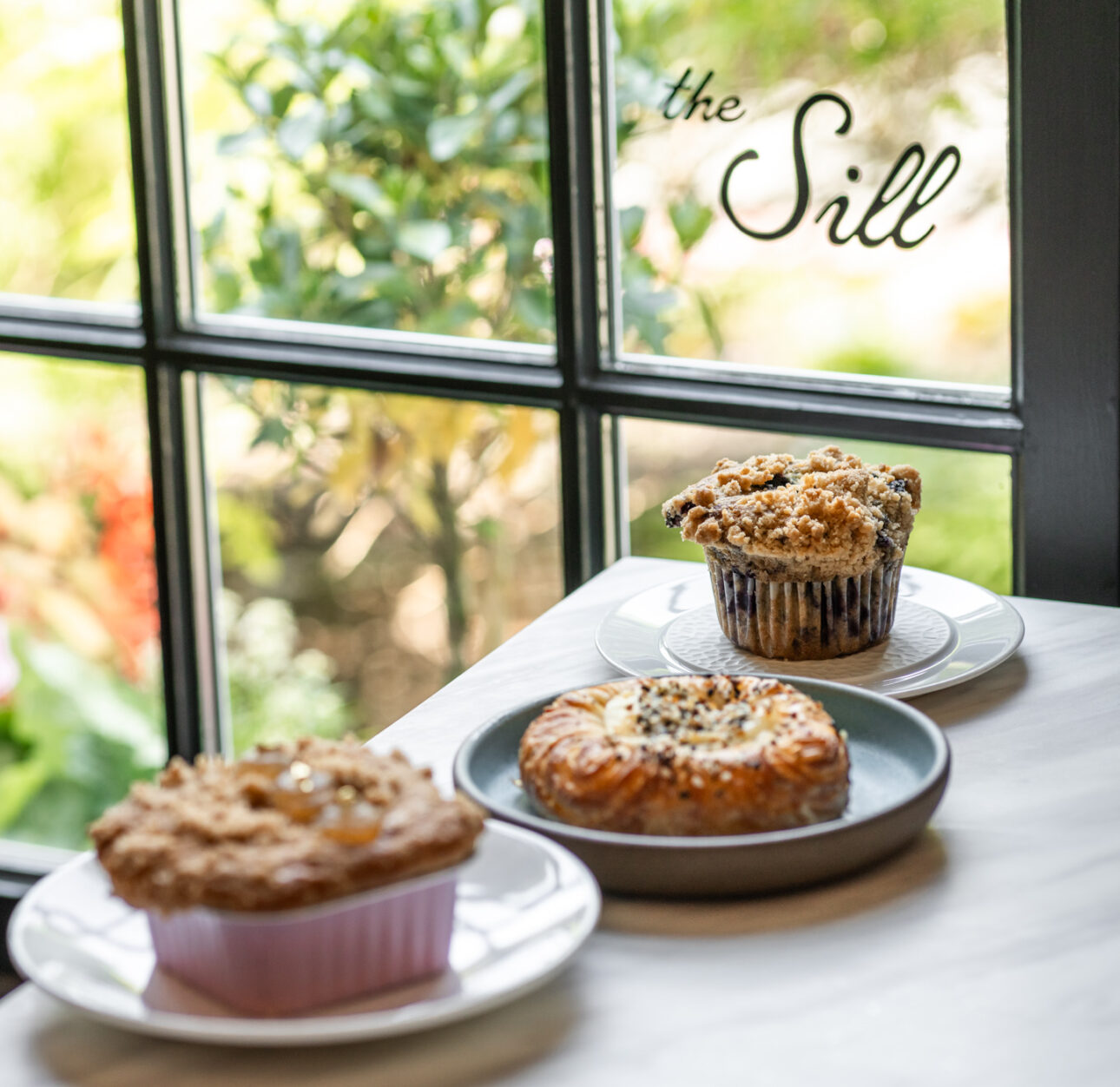 Three pastries sit on plates atop a table by a window with greenery and flowers outside; "the Sill" appears on the glass, evoking the relaxed atmosphere of a resort like Wequassett Resort & Golf Club.