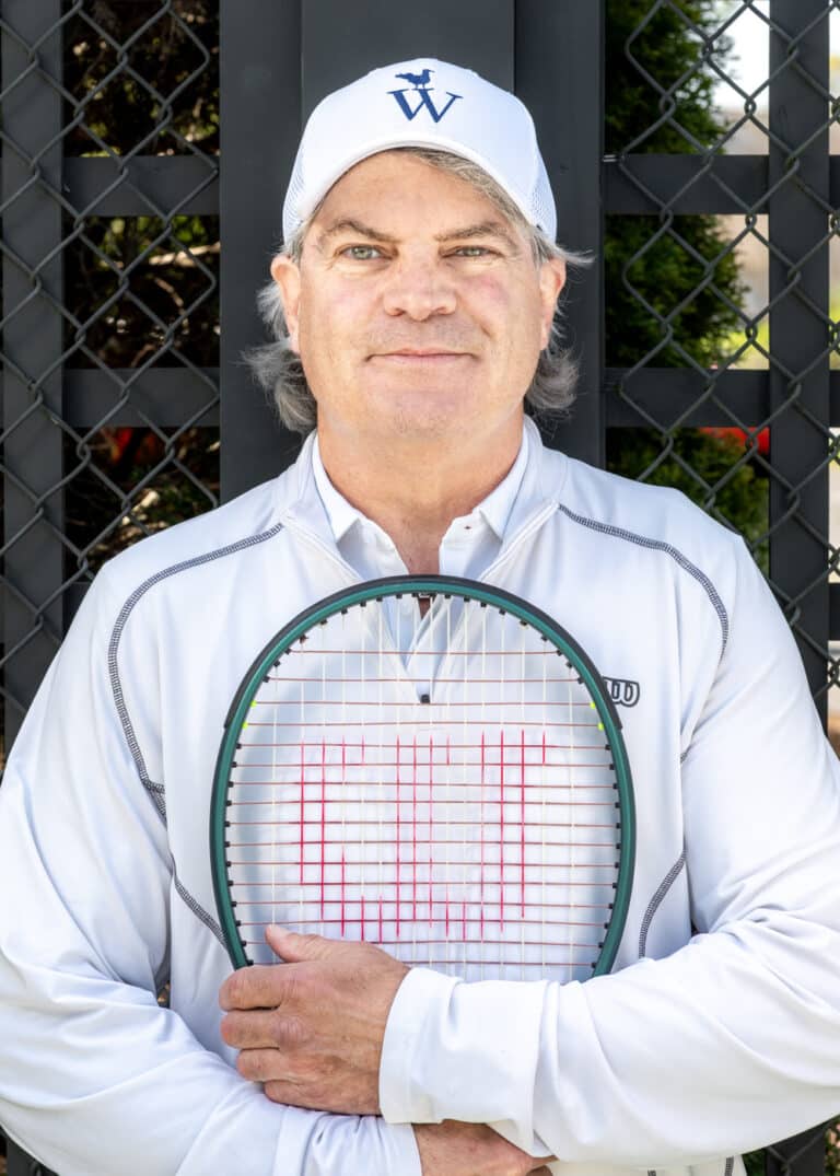 A middle-aged man in a white cap and jacket stands in front of a fence, holding a tennis racket vertically across his chest, ready for a game of Tennis or other Racquet Sports.