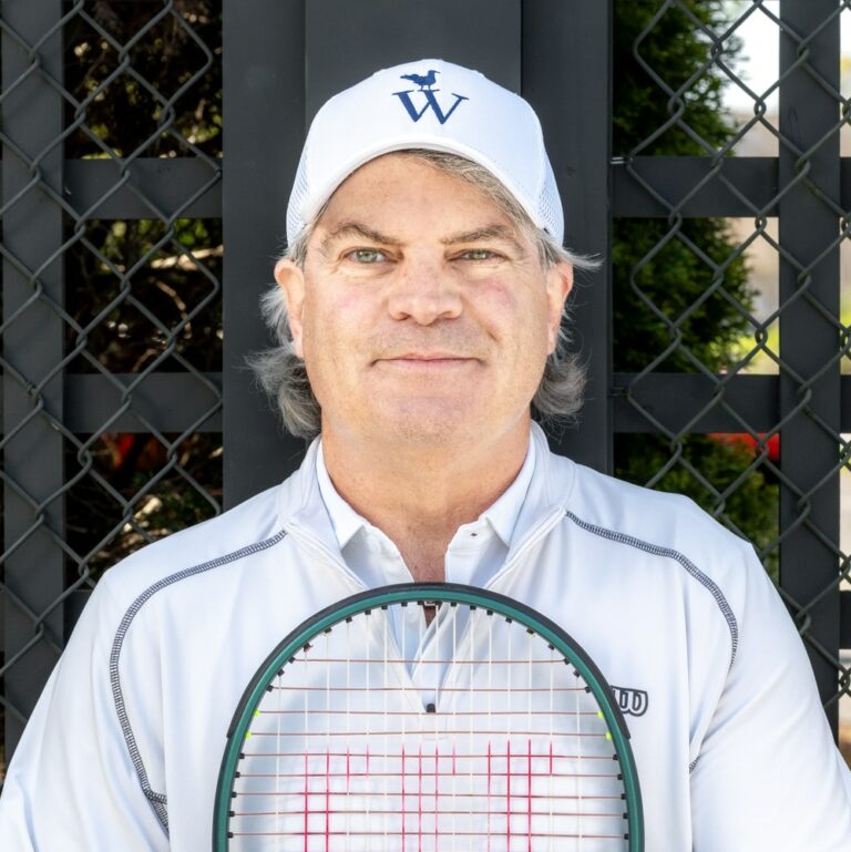 A middle-aged man in a white cap and jacket stands in front of a fence, holding a tennis racket vertically across his chest, ready for a game of Tennis or other Racquet Sports.