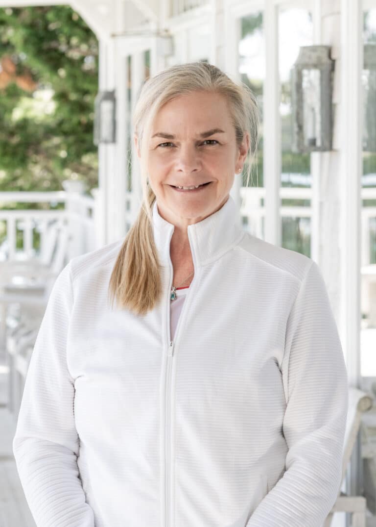A woman with blonde hair in a ponytail, dressed in a white zip-up jacket, stands on a porch with white railings and outdoor furniture—ready for a day of racquet sports.