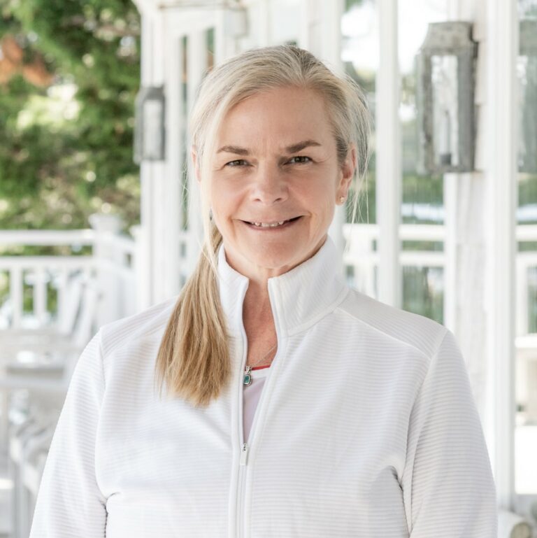 A woman with blonde hair in a ponytail, dressed in a white zip-up jacket, stands on a porch with white railings and outdoor furniture—ready for a day of racquet sports.