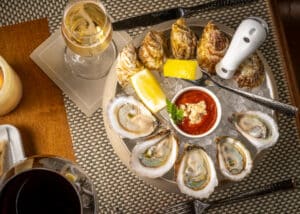 A plate of raw oysters and clams on ice with lemon wedges, cocktail sauce, and a glass of white wine on a woven placemat.