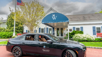 A black BMW sedan is parked in front of a white building with a blue awning labeled "W" at Wequassett; an American flag waves nearby, framed by trees and shrubs in the background.