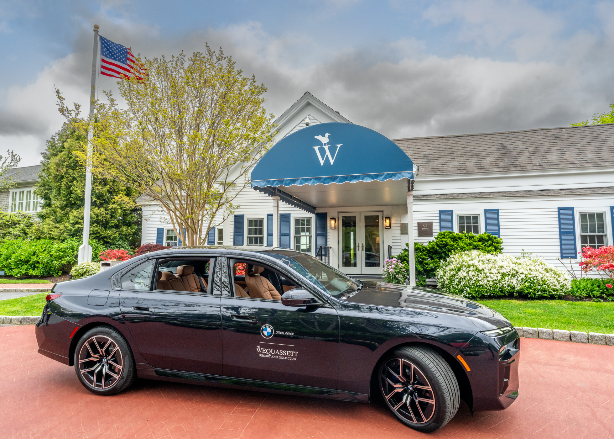 A black BMW sedan is parked in front of a white building with a blue awning labeled "W" at Wequassett; an American flag waves nearby, framed by trees and shrubs in the background.