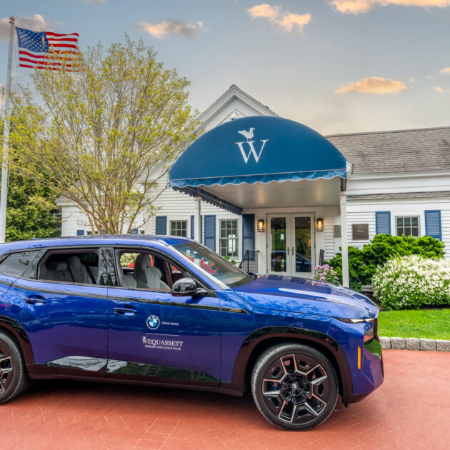 A blue BMW SUV is parked in front of the White Elephant hotel entrance, with an American flag and lush greenery in the background, evoking the elegant vibe found at resorts like Wequassett.