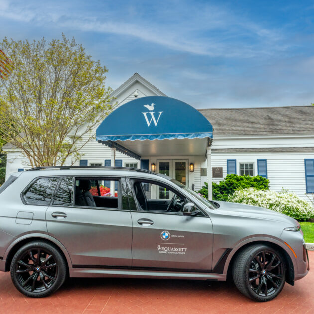 A matte gray BMW SUV is parked in front of a white building with a blue awning and an American flag on a flagpole to the left, evoking the classic charm of Wequassett.