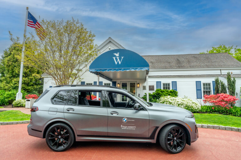 A matte gray BMW SUV is parked in front of a white building with a blue awning and an American flag on a flagpole to the left, evoking the classic charm of Wequassett.