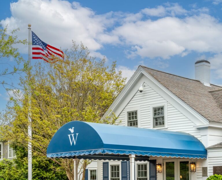 A gray BMW SUV is parked on a red driveway in front of a building with a blue awning and an American flag, offering a glimpse of Coastal Heritage. The sky is partly cloudy.
