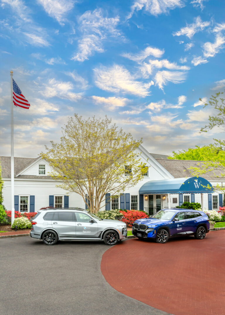 Two SUVs, including a sleek BMW, are parked in front of a white building with a blue awning under a cloudy sky; an American flag waves on a pole to the left.