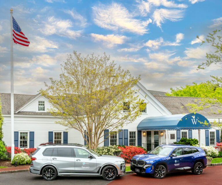 Two SUVs, including a sleek BMW, are parked in front of a white building with a blue awning under a cloudy sky; an American flag waves on a pole to the left.