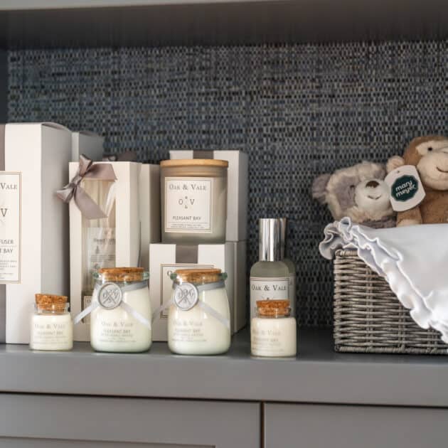 A shelf displays Oak & Vale scented candles, reed diffusers, and sprays—available to shop at Wequassett—alongside a basket with plush toys and a folded white blanket.