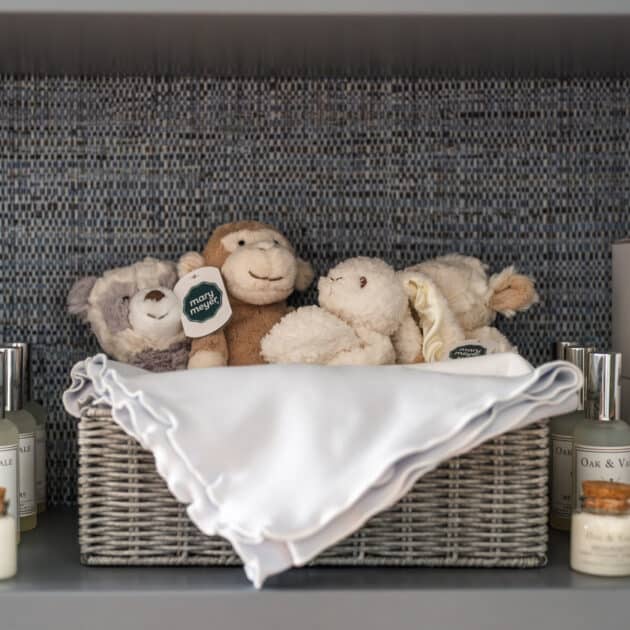 A wicker basket with plush toys and a white cloth is displayed on a shelf at Wequassett, surrounded by candles and bottles of lotion, set against a blue mosaic tile background.