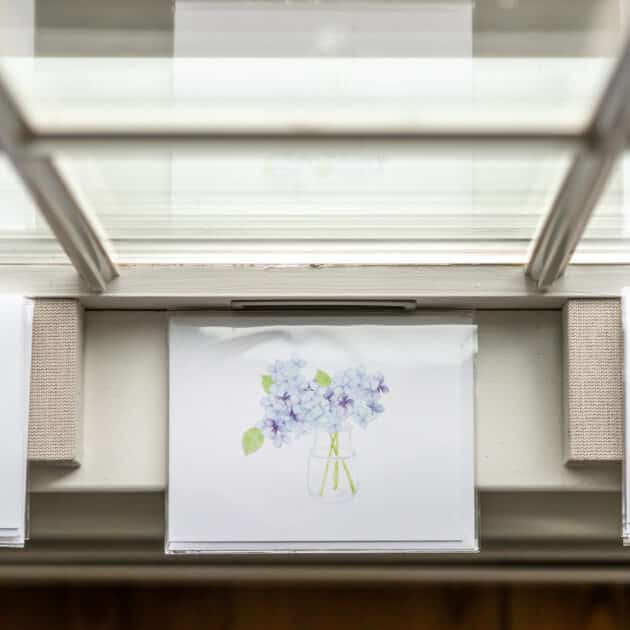 A drawing of light purple flowers in a vase, displayed inside a clear sleeve, is positioned on a windowsill with sunlight streaming through above—capturing the serene coastal charm of Wequassett Resort.