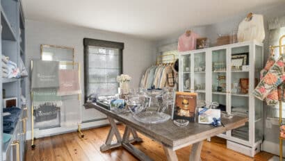 A boutique room displays folded towels, glassware, apparel, and home decor items on shelves and a wooden table, with natural light coming through two windows.