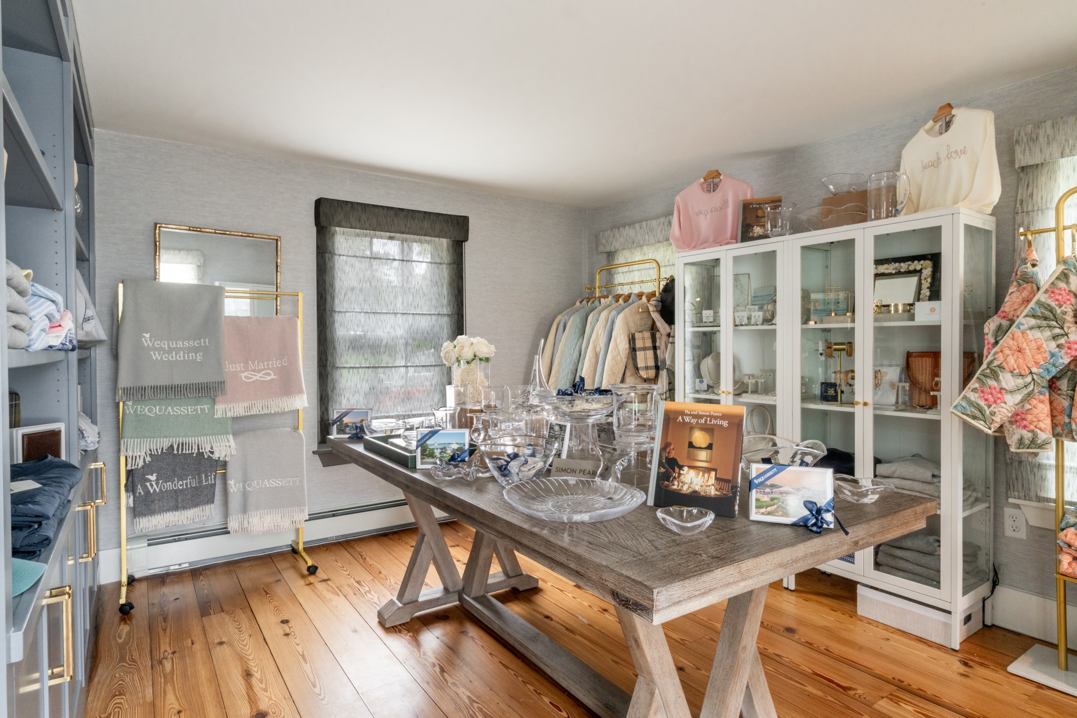 A boutique room displays folded towels, glassware, apparel, and home decor items on shelves and a wooden table, with natural light coming through two windows.