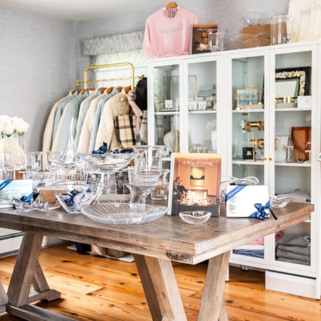A wooden table displays glassware and books in a boutique. Coats and clothing hang on racks, with shelves of accessories and decor in the background.