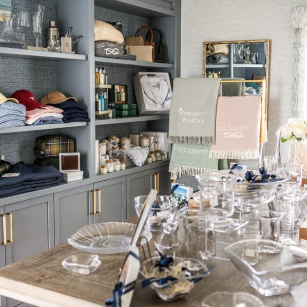 A room with gray built-in shelves holding folded clothes, towels, baskets, and decor items. A wooden table is set with glassware, plates, and small decorative objects.