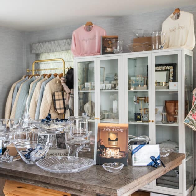 A boutique store display with glassware on a wooden table, clothing on racks, and gift items in a white cabinet against a gray wall.