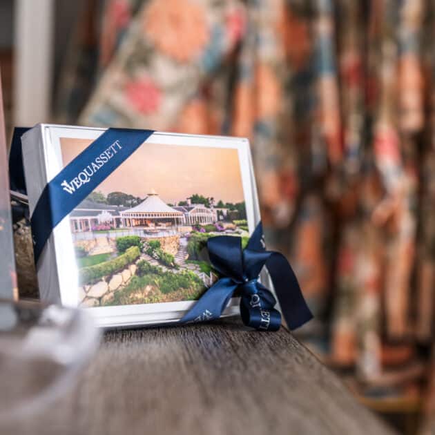 Framed photo of a white building with a blue ribbon labeled “WEDUQUASSET” sits on a wooden surface, accompanied by a glass dish and blurred patterned fabric in the background. Shop Wequassett for more distinctive decor like this.
