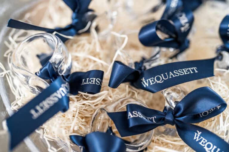 Close-up of clear glass items with navy blue ribbons labeled "WEQUASSETT," arranged on straw-like packing material.