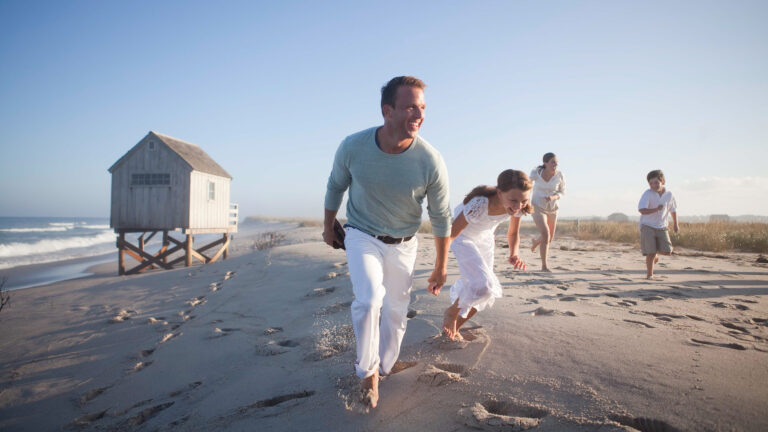 A group of people, including children and adults, walk and run barefoot on a sandy beach near a wooden house on stilts by the ocean at Wequassett Resort and Golf Club.