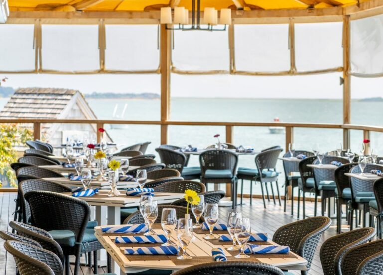 Outdoor restaurant patio with neatly set tables and chairs under a yellow canopy, overlooking water and greenery—perfect for an Annual Fourth of July celebration or summer gala.