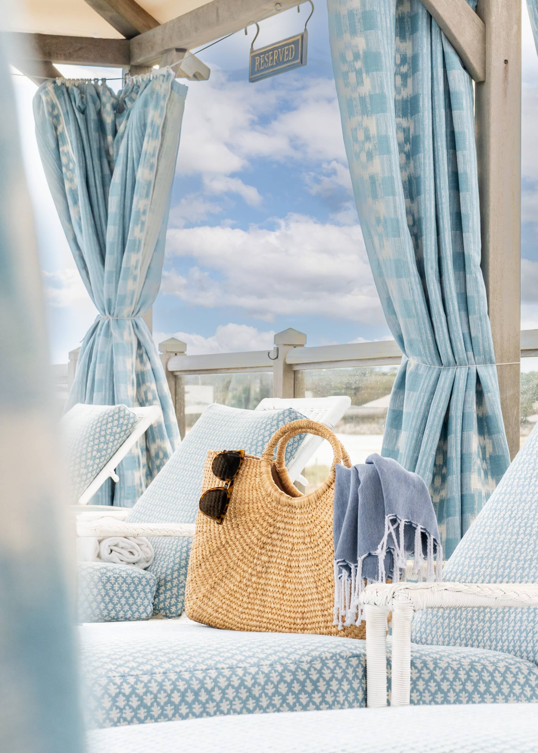 Wicker bag with sunglasses and blue towel on a cushioned lounge chair, set on a deck with blue patterned curtains and a "reserved" sign overhead, capturing the laid-back elegance of La Bella Summer Renewal at Wequassett.