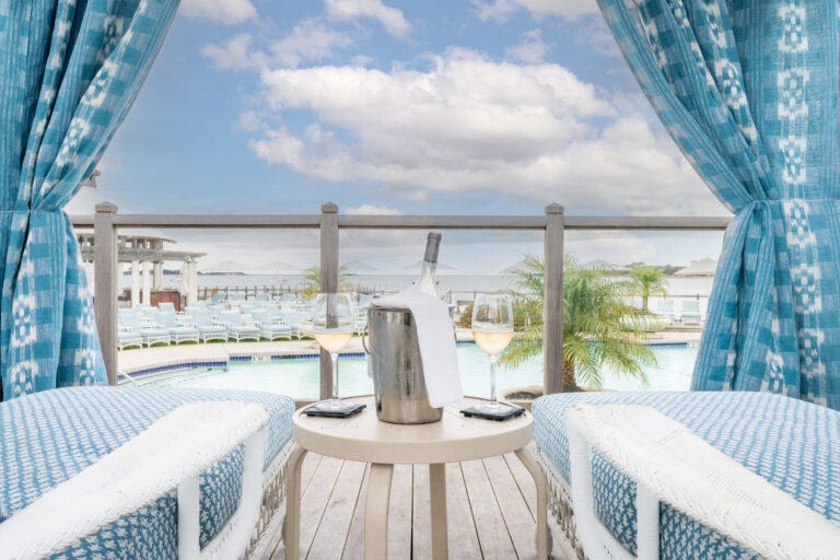 Two lounge chairs with blue and white fabric face a small table with a wine bottle in an ice bucket and two glasses, overlooking the pool and palm trees at Wequassett, a premier Cape Cod hotel, under a cloudy sky.