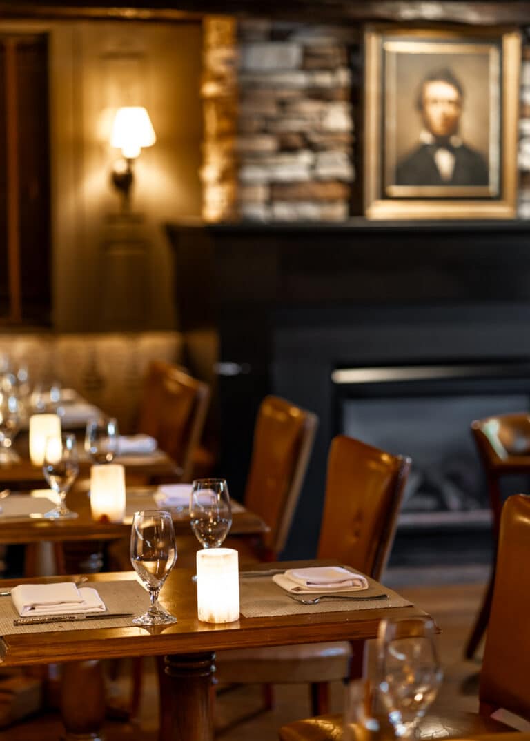 A dimly lit restaurant dining area at Wequassett Resort with set tables, wine glasses, candle-style lights, and a framed portrait above a fireplace.
