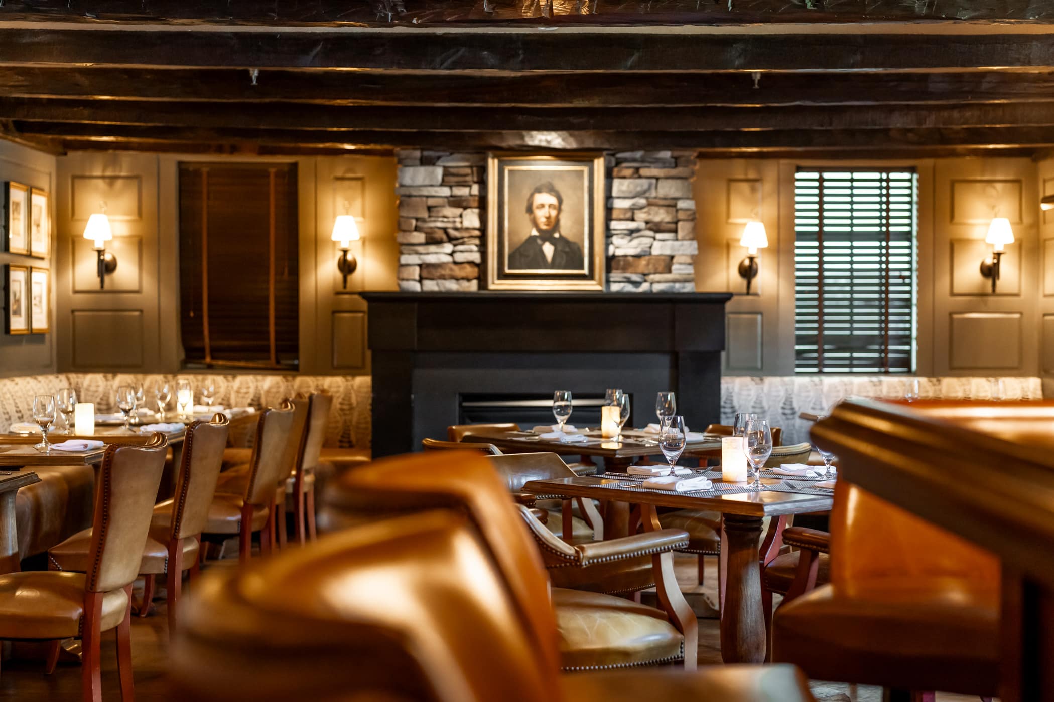 A warmly lit restaurant dining room with leather chairs, set tables, stone fireplace, and a framed portrait above the mantel.
