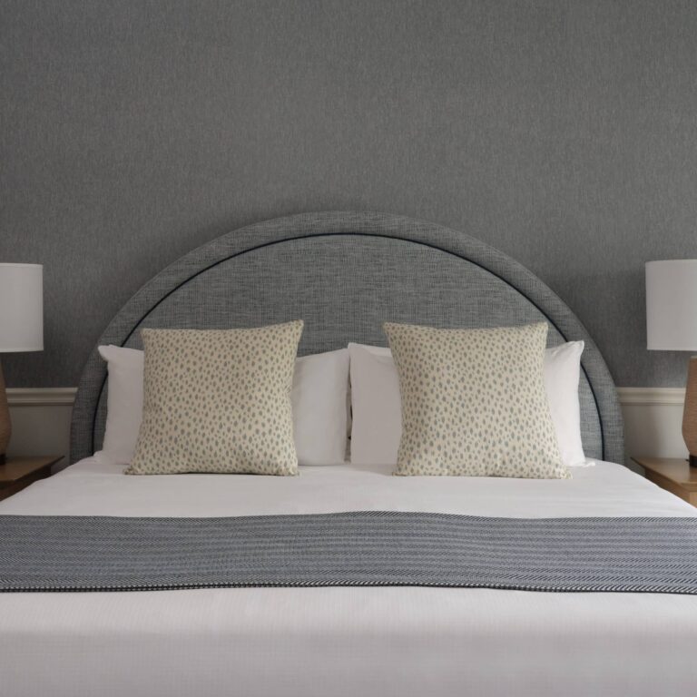 A neatly made bed with two patterned pillows, a grey padded headboard, a grey throw, and two matching lamps on bedside tables against a grey wall—perfect for relaxing after a day at the Cape Cod golf course or nearby Cape Cod hotel.