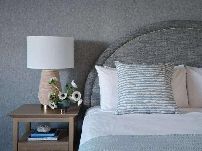 A neatly made bed with a patterned pillow, a wooden nightstand holding a lamp, a vase of white flowers, and stacked books against a gray wall evoke the classic elegance found at 28 Atlantic, an iconic Cape Cod wedding venue.