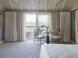 A hotel room at Wequassett Resort and Golf Club features two chairs and a round table near sliding glass doors opening to a balcony with an ocean view and distant sailboats, offering the perfect Cape Cod resort escape.