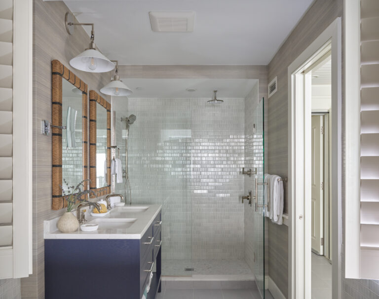 Modern bathroom with a double-sink vanity, large wall mirrors, glass-enclosed shower, and gray tiled walls—echoing the serene luxury found at Wequassett Resort and Golf Club, with natural light streaming through window shutters.