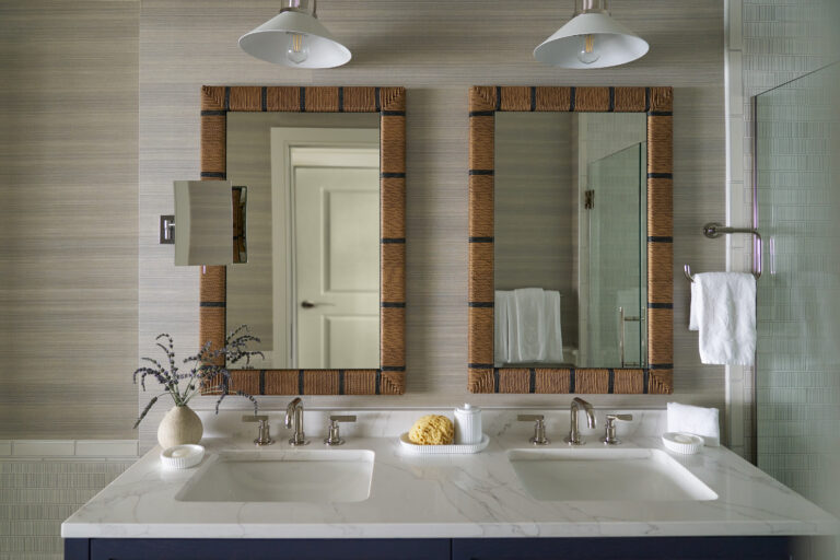 Double bathroom vanity with marble countertop, two sinks, wicker-framed mirrors, wall-mounted lights, and a towel by a glass shower—echoing the coastal luxury of Wequassett Resort and Golf Club or a Cape Cod hotel. A vase and sponge rest on the counter.