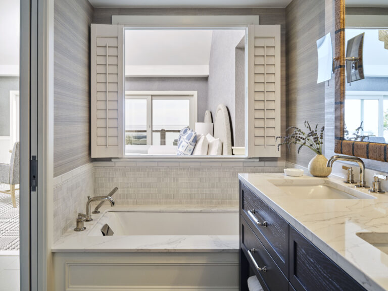 Modern bathroom at Wequassett Resort and Golf Club featuring a marble countertop, built-in bathtub, gold fixtures, and window shutters that open to a bright adjoining bedroom.