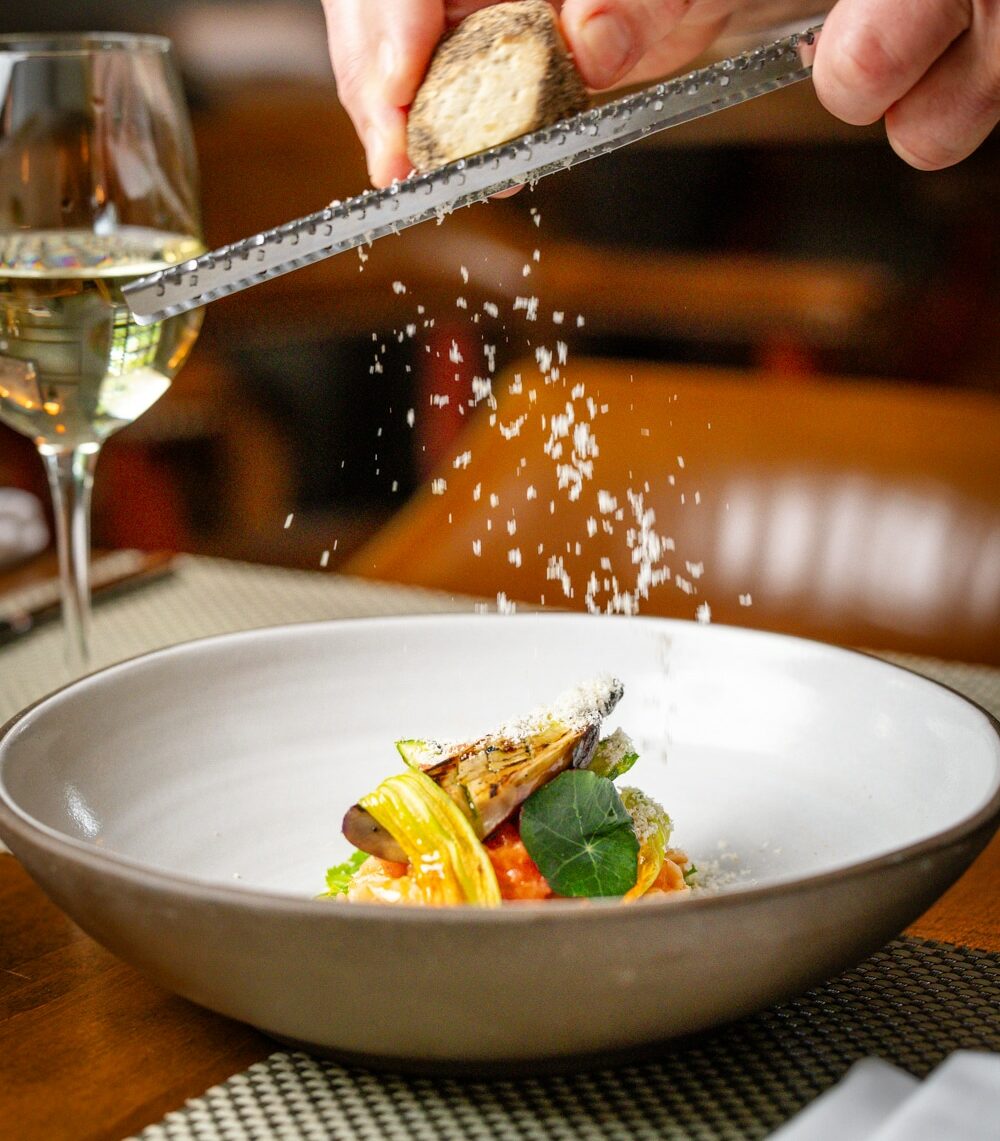 A person grates a truffle over a plated gourmet dish, with a glass of white wine on the table beside it.