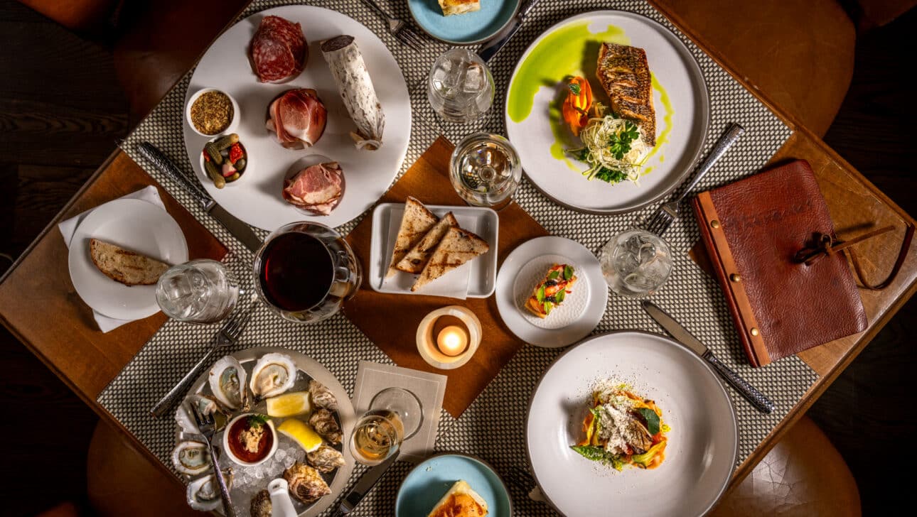Overhead view of a restaurant table set with seafood, charcuterie, grilled fish, bread, wine, a candle, and a menu on woven placemats.