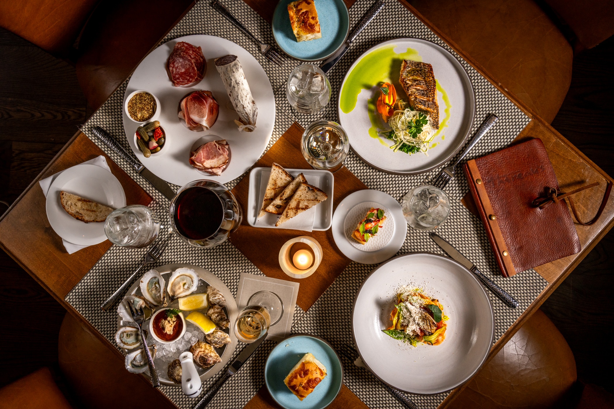 Overhead view of a restaurant table set with seafood, charcuterie, grilled fish, bread, wine, a candle, and a menu on woven placemats.