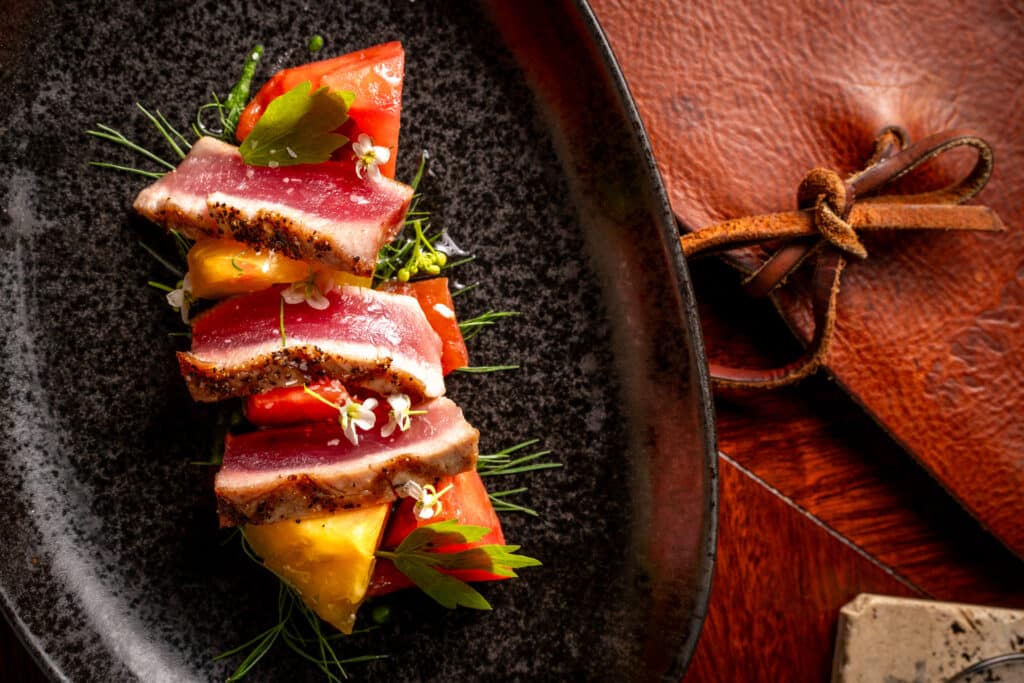 Sliced seared tuna stacked with yellow and red tomatoes, garnished with microgreens, served on a dark plate next to a brown leather pouch.
