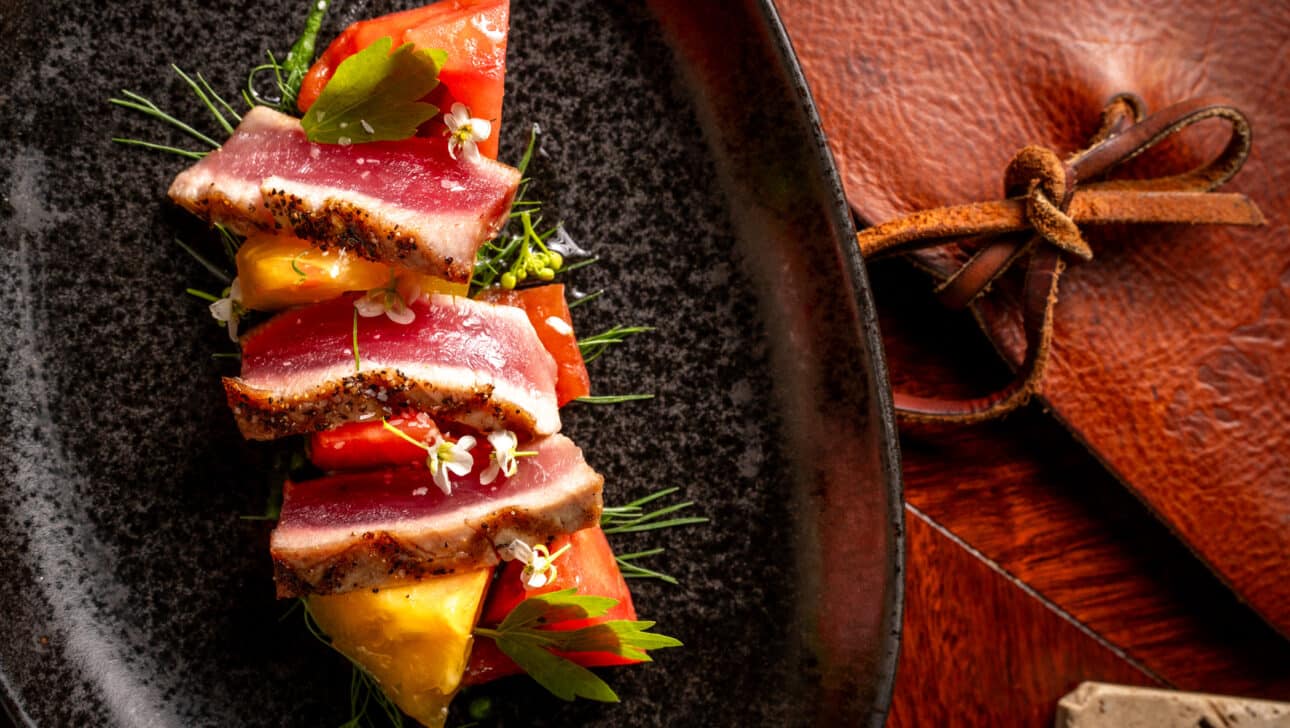 Sliced seared tuna stacked with yellow and red tomatoes, garnished with microgreens, served on a dark plate next to a brown leather pouch.